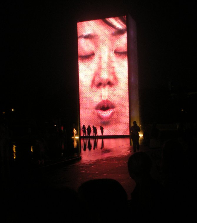 Children and adults alike frolicking after dark in the reflecting pool under one of the two 50-foot glass block towers at Chicago's Millenium Park Crown Fountain. Video images of a broad spectrum of Chicago citizens are projected from the towers and water flows through a water outlet in the screen to give the illusion of water
spouting from their mouths.  
