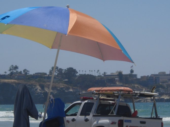 Colorful beach umbrellas offer shade to a passing lifeguard truck at La Jolla Shores beach.
