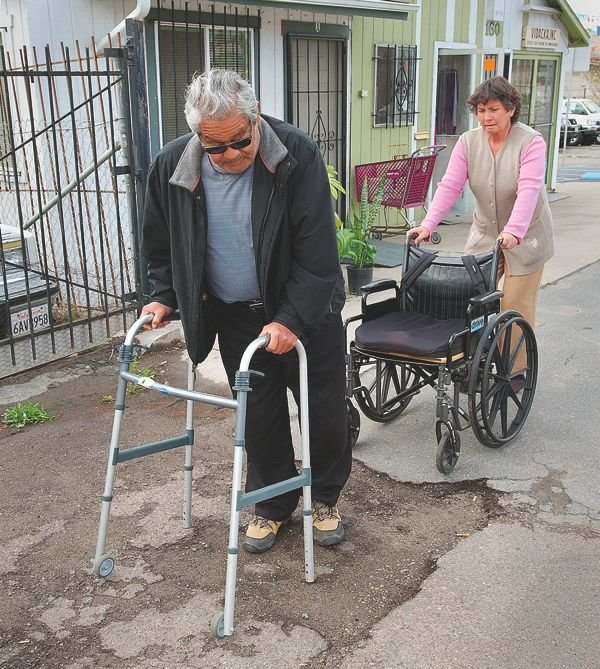 Juan Mariscal struggles to navigate San Ysidro’s crumbling sidewalks using his walker or in his wheelchair (his wife Irma behind him).