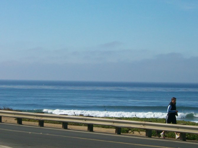 A jogger runs along Sunset Cliffs Blvd. with surfers in the background.