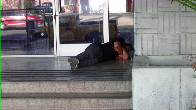 Homeless individuals sleep near the entrance and on the side of the building.