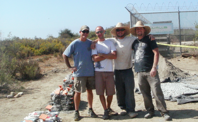 Two volunteers stand with Waylon Matson and Steven Wright (3rd and 4th from left) of 4 Walls International. Soda-bottle-filled bench bases on the left.
