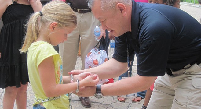 A U.S. Marshals official shows a girl how restraining cuffs work. Local defense attorneys aren’t happy that all of their clients must wear them in federal court.