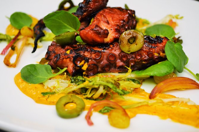 Charred octopus from Sea & Smoke served on a bed of ancho chili romesco, with wilted frisee, golden raisins, and castelvetrano olives and fresh lemon juice.