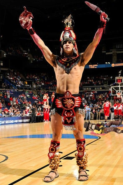 San Diego State mascot “Monty” Montezuma bears aloft the freshly harvested heart of Cowboy Joe [in background, bleeding and dead], mascot for the Aztecs' Mountain West Conference rival the University of Wyoming. In keeping with the ancient Aztec tradition of “feeding the gods” with sacrificed flesh, the heart is flash-fried and then fed to members of the men’s basketball team immediately prior to tip-off. “The sun god Tezcatlipoca needed nourishment to raise the sun each morning, and [San Diego State center Valentine] Izundu needs nourishment to jump up and touch the orange orb lofted toward heaven by the referee,” explains team trainer Verne Beefcheeks. “And just as the Aztecs would sometimes go to war precisely to obtain victims for sacrifice, so our boys go up against rivals like the Cowboys to ensure the health and prosperity of our entire athletic program. It all makes sense.”