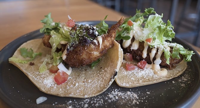 A pair of fried lobster-tail tacos topped by lettuce, tomato, onion, a dusting of cotija, and a sweet-chile aioli