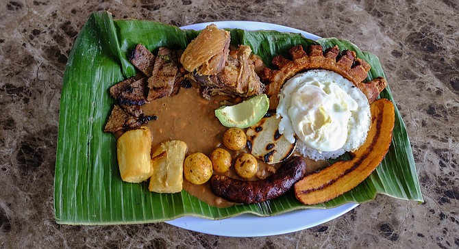 Clockwise from the top left corner: carne asada, fried chicken, crispy pork belly, fried plantain, sausage, and fried cassava. Fried egg, rice, arepa, potatoes, beans, and avocado in the center.