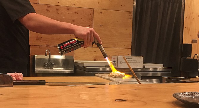 A sushi chef sears several pieces of black cod with a blowtorch.