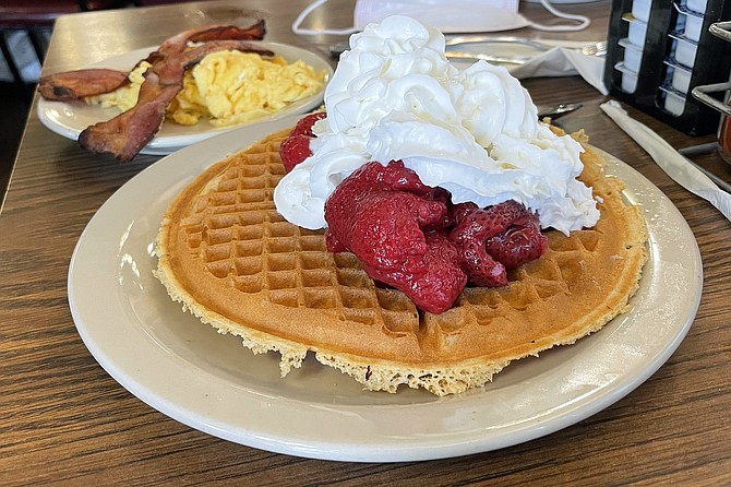 Preserved strawberries and whipped cream top a large waffle, with a side of eggs and bacon
