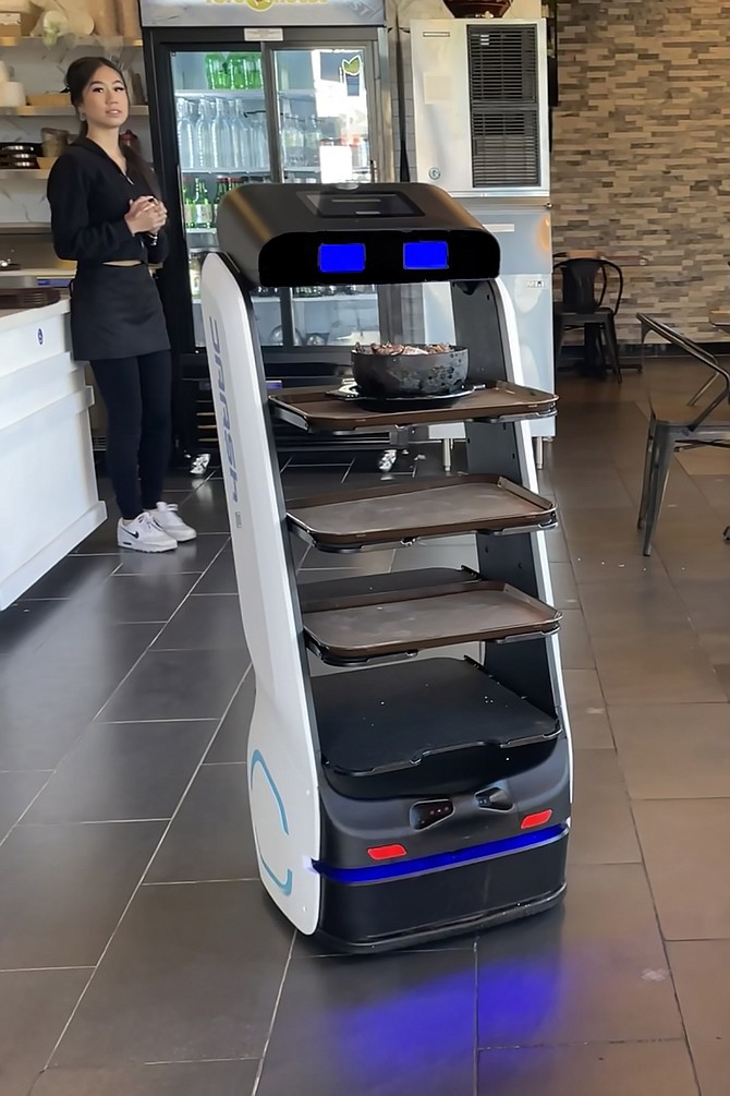 The robot brings the hot dish to the table, while the waitress stands by to transfer it to the table.